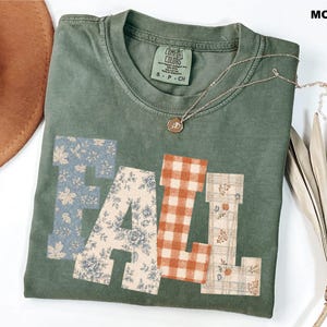 May include: A moss green short-sleeve t-shirt featuring the word FALL in large block letters. Each letter has a different pattern: blue leaves, cream floral, orange gingham, and a plaid print with small pumpkins. The shirt has a crew neckline.