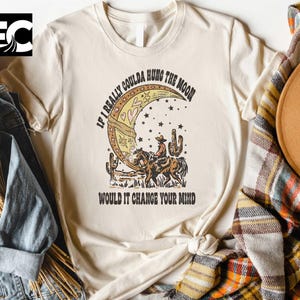 May include: Cream-colored t-shirt featuring a vintage-style graphic of a cowboy riding a horse in front of a crescent moon. The text reads, "IF I REALLY COULDA HUNG THE MOON WOULD IT CHANGE YOUR MIND". The design includes stars and cacti.