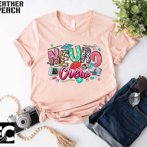 a pink t-shirt with a colorful graphic design, a pair of blue jeans, a camera, and some accessories such as a plant and sunglasses.