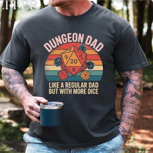 May include: Dark gray t-shirt with a colorful graphic featuring a D20 die and the text "DUNGEON DAD LIKE A REGULAR DAD BUT WITH MORE DICE". The design includes a retro sunset and other dice.