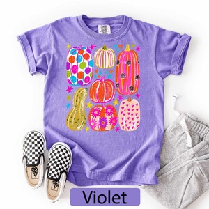 May include: A violet short-sleeve t-shirt featuring a graphic print of various pumpkins in bright colors including pink, orange, yellow, and blue, decorated with patterns like polka dots, stripes, flowers, and hearts, alongside small star shapes.