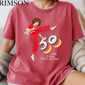 A red t-shirt with a graphic design depicting a woman in a red outfit with the number "50" on it. The text below the graphic says "I Like To Kick Stretch And Rick!"
