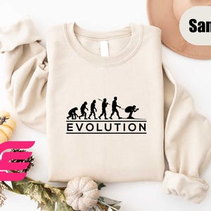 May include: A beige sweatshirt with a black graphic of the evolution of man, ending with a skier. The graphic is labeled "EVOLUTION".
