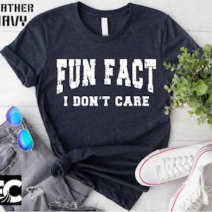 May include: Heather navy t-shirt with the text "FUN FACT I DON'T CARE" in white block letters. The shirt is styled with sunglasses, jeans, and white sneakers. The shirt is a casual, everyday wear item.