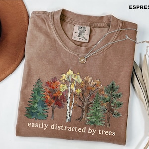 May include: An espresso-colored short-sleeve t-shirt featuring a graphic of various trees in autumn colors. The text "easily distracted by trees" is printed in white below the graphic. The shirt has a crew neckline and a Comfort Colors brand label.