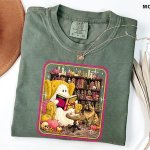 May include: A moss green t-shirt featuring a graphic of a ghost sitting in a yellow armchair reading a book titled Dungeon Crawler Carl, with a cat nearby, a bookshelf, pumpkins, and a skull. The shirt has a Comfort Colors label at the neck.