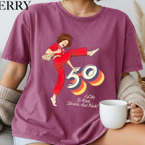 A person wearing a purple t-shirt with a graphic design of a woman in red pants and a white top, along with the text "I like to kick, stretch, and kick!"