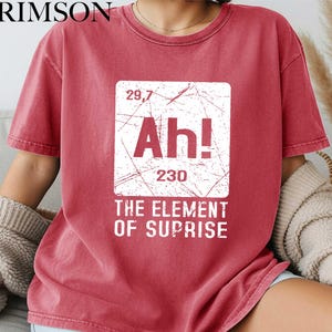May include: Crimson red t-shirt featuring a distressed white graphic. The graphic reads "Ah!" with numbers and the phrase "THE ELEMENT OF SUPRISE". The shirt has a crew neck and short sleeves.
