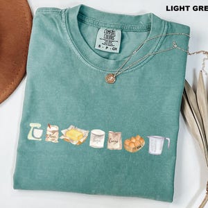 May include: A light green short-sleeve t-shirt with a crew neckline and a graphic print featuring a stand mixer, a bag labeled flour, a stick of butter, a container labeled sugar, a bag labeled cacao, a carton of eggs, and a measuring cup.