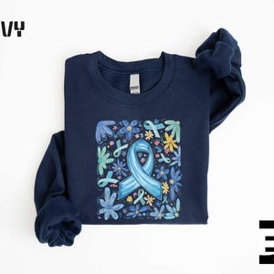 May include: Navy blue sweatshirt featuring a floral design with a light blue ribbon and flowers. The word "NAVY" is printed in bold, white letters at the top left. The sweatshirt has a crew neck and long sleeves. The design includes various blue, yellow, and green flowers.