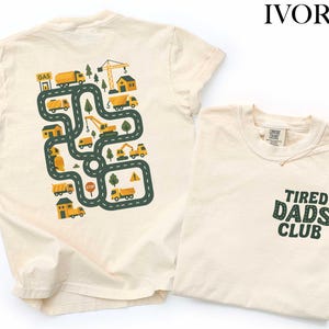 May include: Ivory-colored t-shirts. One shirt features a road map design with trucks, construction vehicles, and buildings. The other shirt has "Tired Dads Club" printed on the pocket in green letters. Both shirts are made of a soft material.