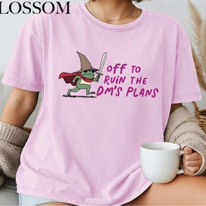 May include: Light pink t-shirt with the text "OFF TO RUIN THE DM'S PLANS" and a cartoon frog wearing a wizard hat and cape, holding a sword. The word "BLOSSOM" is printed at the top left.