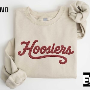 May include: A sand-colored sweatshirt with the word "Hoosiers" embroidered in a burgundy script font. The sweatshirt has a crew neck and long sleeves, with the word "SAND" printed in black block letters in the upper left corner.