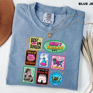 May include: A blue jean colored t-shirt featuring a graphic print of multiple rectangular and circular badges with various illustrations and text including Best in Dungeon, New Achievement, This is an Outrage, Mongo is Appalled, and Princess Donut.