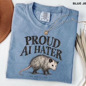 May include: A blue crew neck t-shirt featuring the text PROUD AI HATER in a dark, distressed font above an illustration of an opossum. Below the opossum, the text reads made poorly by a human. The shirt has a Comfort Colors brand neck label.