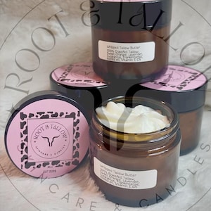 May include: Two glass jars of whipped tallow butter with pink and black labels. The labels have a cow skull logo and the text "Root & Tallow Skincare & Candles Est 2023". The jars are filled with a white, creamy substance.