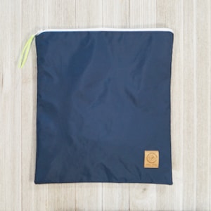 May include: A dark blue zippered pouch with a white zipper and a small brown leather patch with a logo. The pouch is lying flat on a light wood surface.