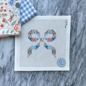 Può includere: Una tela quadrata per ricamo a mezzo punto con un disegno centrale a fiocco composto da piccoli motivi geometrici multicolori nelle tonalità del blu, rosso e verde. Un piccolo adesivo circolare con la lettera B è applicato nell'angolo in basso a destra.