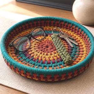 May include: A round, multi-colored crochet tray with a teal border. The tray holds black-framed glasses, keys, and a green key fob. The tray's interior features a vibrant pattern of orange, yellow, and teal.