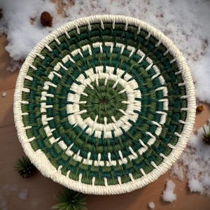 May include: A round, woven basket with a spiral design. The basket features alternating bands of dark green and white, with a cream-colored border. The basket is approximately 6 inches in diameter.
