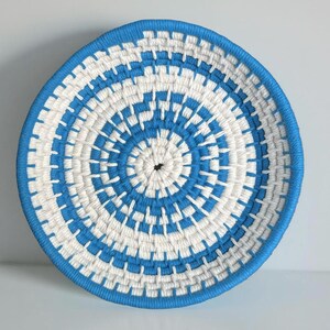 May include: A round, woven basket with a blue and white spiral design. The basket has a blue rim and a central hole. The pattern is a repeating geometric design. The basket is likely handmade and suitable for home decor.