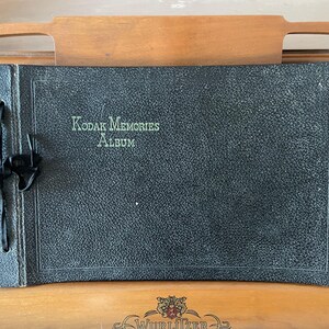 May include: A rectangular black photo album with a textured cover featuring the text Kodak Memories Album in light-colored lettering. The album is bound on the left side with a black ribbon tied in a bow through two punched holes in the cover.