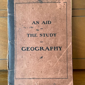 May include: A vintage book with a weathered, light brown paper cover and a black cloth spine. The front cover features the printed text AN AID IN THE STUDY OF GEOGRAPHY in black capital letters, with a small decorative flourish at the bottom.