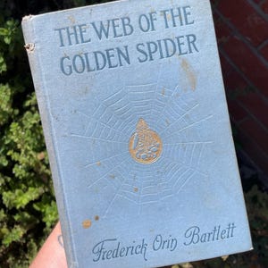 May include: A light blue hardcover book titled The Web of the Golden Spider by Frederick Orin Bartlett. The front cover features a debossed spider web design with a central gold circular emblem depicting a seated figure, showing signs of wear.