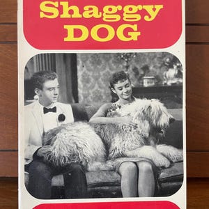 May include: A vintage paperback book titled The Shaggy Dog with a red border and yellow text. The cover features a black and white photograph of a person in a tuxedo and a person in a dress sitting on a sofa with a large, long-haired dog.