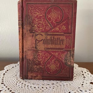 May include: A vintage hardcover book with a deep red cover featuring intricate gold-colored filigree and floral patterns. The title 'Palmbl&auml;tter' is embossed in a decorative gothic-style font across the center of the front cover.