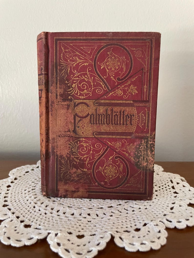 May include: A vintage hardcover book with a deep red cover featuring intricate gold-colored filigree and floral patterns. The title 'Palmbl&auml;tter' is embossed in a decorative gothic-style font across the center of the front cover.