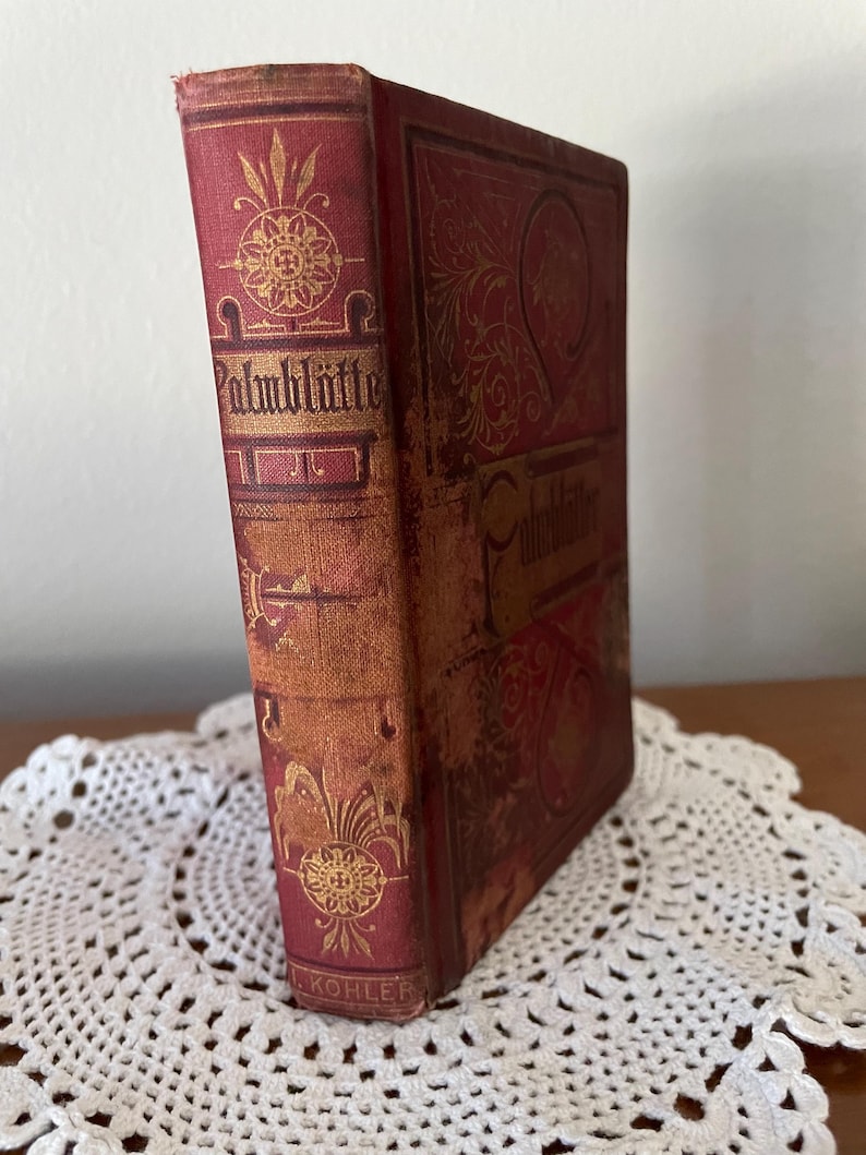 May include: A red hardcover book titled Palmbl&auml;tter with gold embossed floral and geometric patterns on the spine and front cover. The spine features the name L. KOHLER at the bottom. The book has a worn texture and stands upright.