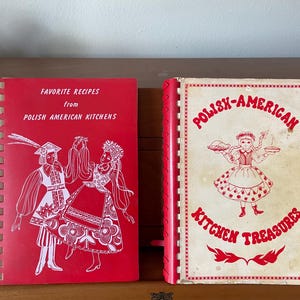 May include: Two spiral-bound cookbooks. The left book is red with white text reading Favorite Recipes from Polish American Kitchens and a white illustration of two dancers. The right book is cream with red text reading Polish-American Kitchen Treasures.