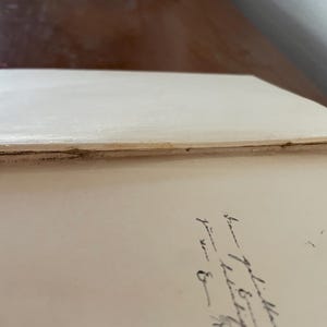 May include: The interior of an open hardcover book with cream-colored endpapers. The spine hinge shows visible wear, fraying, and separation of the binding material. On the right-hand page, there is a handwritten inscription in black ink.