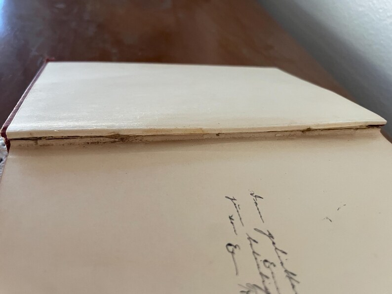 May include: The interior of an open hardcover book with cream-colored endpapers. The spine hinge shows visible wear, fraying, and separation of the binding material. On the right-hand page, there is a handwritten inscription in black ink.