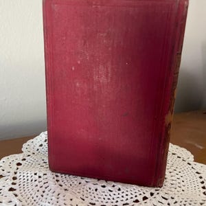 May include: A rectangular hardcover book with a solid deep red fabric cover. The front cover features a subtle embossed border frame design. The spine of the book shows signs of wear with visible fraying along the edges and corners of the binding.