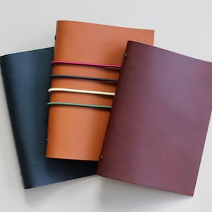 May include: Three leather journals in shades of navy, tan, and burgundy. The tan journal is secured with colorful elastic bands. The journals are arranged on a light-colored surface, showcasing their smooth textures and simple design.