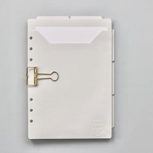 May include: A white, textured paper notebook with a gold-colored binder clip. The notebook has a pocket and tabbed dividers. The notebook is on a light gray background. The notebook has a logo in the bottom right corner.