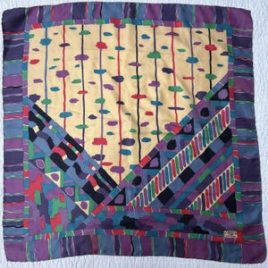 May include: Square silk scarf with a geometric design. The scarf features a cream-colored center with vertical lines and colorful cloud-like shapes. The border is a mix of purple, blue, and green geometric patterns. The scarf has a label that says "Made in England."