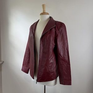 May include: A burgundy leather jacket with a zip-up front is displayed on a mannequin. The jacket has a collar and long sleeves. The mannequin is on a metal stand against a white background. The jacket appears to be in good condition.