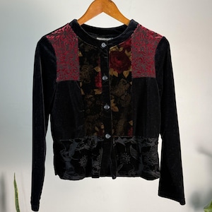 May include: Black velvet jacket with long sleeves and a round collar. The jacket features panels of red floral brocade and a central panel with a floral pattern. The bottom hem has a black floral design and is buttoned down the front.
