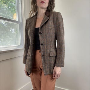 May include: A brown plaid blazer featuring a dark brown velvet collar, three front buttons, and two flap pockets. The jacket is worn over a black top and orange trousers. The plaid pattern consists of intersecting lines in shades of brown and blue.