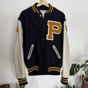 May include: Black varsity jacket with cream leather sleeves. It has a large gold "P" on the chest and gold and black striped trim on the collar, cuffs, and hem. A classic vintage style.