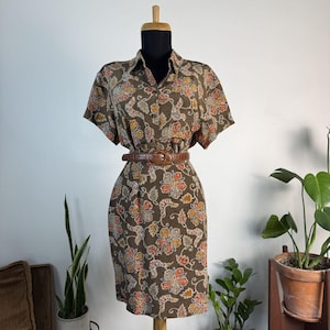 May include: A short-sleeved dress in a dark brown hue featuring a repeating floral pattern in shades of orange, yellow, and white. The dress has a collared neckline, a button-down front, and is cinched at the waist with a brown textured belt.