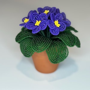May include: A decorative potted plant sculpture crafted from small beads. The plant features deep purple petals with bright yellow centers and textured dark green leaves, all arranged in a simple terracotta-colored clay pot.