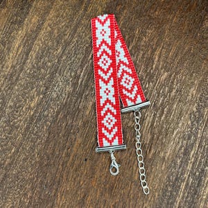 May include: A beaded bracelet with a red and white geometric pattern. The bracelet has a silver-colored clasp and chain extender. The design features repeating diamond and cross-like shapes. The bracelet is displayed on a wooden surface.