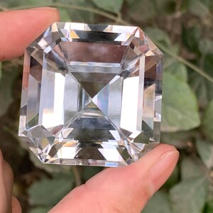 May include: A large, clear, faceted gemstone with a square emerald cut. The gem is held between fingers, showcasing its brilliant clarity and multiple facets. The background features blurred green foliage.