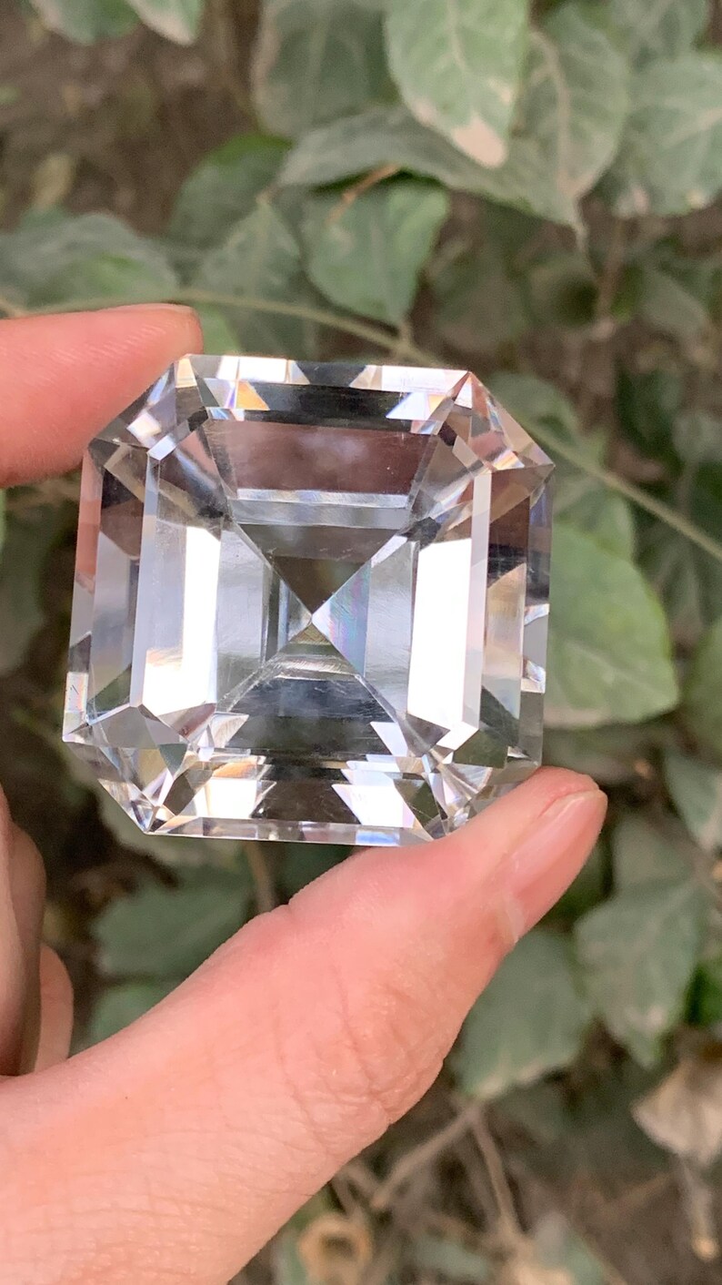 May include: A large, clear, faceted gemstone with a square emerald cut. The gem is held between fingers, showcasing its brilliant clarity and multiple facets. The background features blurred green foliage.