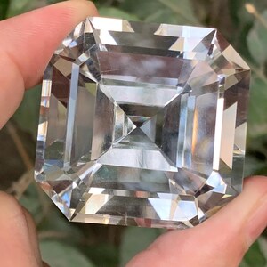 May include: A large, clear, square-cut gemstone with multiple facets, held between fingers. The gem reflects light, creating a brilliant display. The gemstone is a square shape with a smaller square in the center.