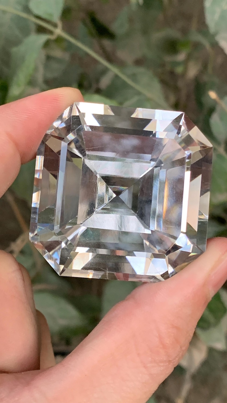 May include: A large, clear, square-cut gemstone with multiple facets, held between fingers. The gem reflects light, creating a brilliant display. The gemstone is a square shape with a smaller square in the center.